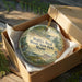 A round ornament in a wooden box, featuring a scenic landscape with a lake and trees, is displayed on a wooden surface.