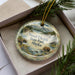 A decorative ornament in a box, featuring a scenic landscape with moose, bears, and waterfalls, with the text "Wood-Tikik State Park, Alaska" printed on it.