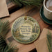 A round ornament with a painting of a historical site, specifically Wormsloe Historic Site, is displayed on a wooden surface alongside a cup of coffee and some pine branches.