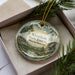 A round ornament with a scenic forest landscape, featuring pine trees, a river, and a rocky outcropping, is displayed in a box.