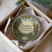 A round ornament in a box, featuring a scenic landscape with pine trees, a lake, and a sign that reads "Wintham State Forest, Massachusetts".