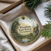 A round ornament with a scenic forest landscape, featuring pine trees, a river, and mountains, is displayed in a box with pine branches.