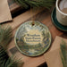 A circular ornament with a scenic forest landscape hangs on a wooden surface, accompanied by a cup of coffee and some pine branches.