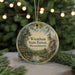 A circular ornament with a scenic forest landscape and directional signs, hanging in front of a blurred Christmas tree with lights.