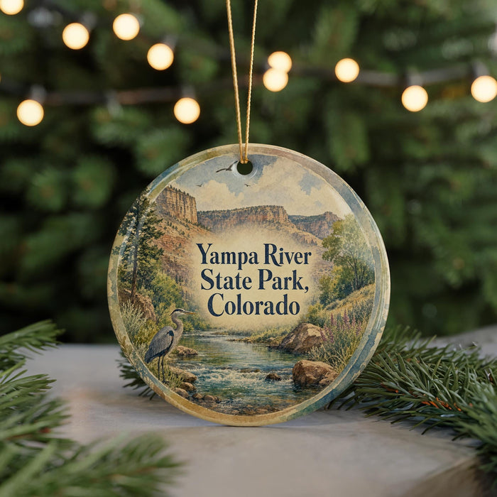 A round ornament with a painting of a river and mountains, featuring a crane bird, hangs in front of a blurred background with Christmas lights.