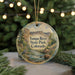 A round ornament with a painting of a river and mountains, featuring a crane bird, hangs in front of a blurred background with Christmas lights.