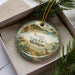 A decorative ornament in a box, featuring a scenic landscape with a bird, pine tree, and deer.