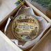 A circular ornament with a painting of a historical building and a desert landscape, placed in a box with a white tissue paper lining.