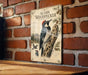a vintage-style metal sign with an illustration of a woodpecker on a tree branch, mounted on a brick wall.