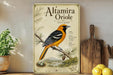 a vintage-style poster of an orange and black bird, specifically the Alamira Oriole, perched on a branch. The poster is displayed on a shelf next to a wooden cutting board and a potted plant.