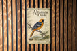 a vintage-style metal plaque with an illustration of a bird perched on a branch, set against a wooden background.