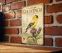 A vintage-style metal sign featuring an illustration of a yellow American Goldfinch perched on a thistle plant, mounted on a brick wall.