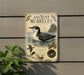 a metal sign with an illustration of a bird, likely a seagull, swimming in the water. The sign is mounted on a wall and is surrounded by greenery, including a plant to the left.