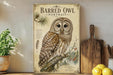 a framed illustration of a barred owl, a type of owl, on a branch with pine needles and berries. The owl is depicted in a detailed and realistic style, showcasing its distinctive features such as its large eyes, pointed beak, and striped plumage. The framed artwork is displayed on a shelf, surrounded by various kitchen utensils and decor items, including a potted plant, a cutting board, and a bowl of fruit.