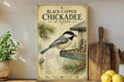 A vintage-style poster of a black-capped chickadee, featuring a detailed illustration of the bird perched on a bird feeder, surrounded by pine cones and pine needles. The poster is displayed on a wooden shelf, with a potted plant and a wooden cutting board visible in the background.