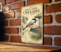 A vintage-style metal sign with a black and white illustration of a chickadee perched on a bird feeder, mounted on a brick wall.