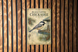 A vintage-style poster of a black-capped chickadee bird is displayed on a wooden wall, with pine cones and pine needles in the background.