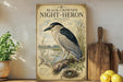 a vintage-style poster of a black-crowned night-heron, a species of bird, perched on a branch with a nest nearby. The poster is displayed on a shelf next to a wooden cutting board and a potted plant.