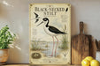 a vintage-style illustration of a black-necked stilt, a type of wading bird, standing on a wooden surface. The illustration includes a detailed depiction of the bird's appearance, habitat, and behavior. The background of the illustration depicts a natural setting with water, plants, and a bird's nest. The image also includes text that provides information about the black-necked stilt, such as its size, habitat, and behavior.