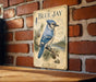 A vintage-style metal sign featuring a blue jay bird on a branch, mounted on a brick wall.