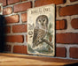 a vintage-style metal sign with an illustration of a Boreal Owl perched on a branch, surrounded by pine cones and pine trees. The sign is mounted on a brick wall, and there is a wooden table below it.