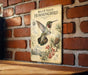 a vintage-style metal sign with a detailed illustration of a hummingbird, mounted on a brick wall.