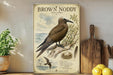 A vintage-style poster of a brown nody bird, featuring illustrations of the bird, its nest, and other elements, is displayed on a shelf next to a wooden cutting board and a potted plant.