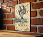 A vintage-style metal sign featuring an illustration of a pelican diving into the water, mounted on a brick wall.