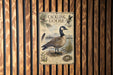 A vintage-style metal sign featuring a large illustration of a Canadian goose, surrounded by various natural elements such as pine trees, grass, and a bird's nest.