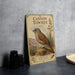 A vintage-style metal plaque depicting a bird, likely a canyon towhee, standing on a rock in a desert landscape.