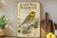a vintage-style poster of a yellow and black bird, specifically the Cape May Warbler, perched on a pine branch. The poster is displayed on a shelf next to a wooden cutting board and a potted plant.