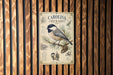 A metal sign with a bird illustration and text is mounted on a wooden wall.