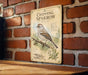 a vintage-style metal sign with a detailed illustration of a chipping sparrow perched on a branch, mounted on a brick wall.