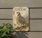 a vintage-style metal plaque with an illustration of a bird, likely a chukar, perched on a rocky outcropping. The plaque is mounted on a wall, and there is a plant visible to the left of the plaque.