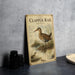 a vintage-style metal plaque or poster with a detailed illustration of a Clapper Rail, a type of wading bird. The plaque is placed on a gray countertop, and there are some black camera lenses and a white bowl visible in the background.