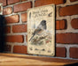 a vintage-style metal sign with a detailed illustration of a bird, likely a sparrow, perched on a branch with berries and pine cones. The sign is mounted on a brick wall, and there is a wooden table below it.