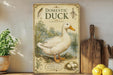 a vintage-style poster of a domestic duck, with illustrations of a duck, a pond, and a nest with eggs, displayed on a shelf next to a wooden cutting board and some potted plants.
