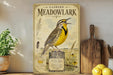 a vintage-style poster of a yellow bird perched on a wooden post, surrounded by illustrations of plants and a map, all set against a white wall.