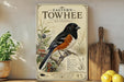 a vintage-style poster of a Towhee bird, which is a small songbird with a black and orange plumage. The poster is displayed on a shelf next to a wooden cutting board and a potted plant.