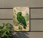 A vintage-style metal plaque featuring a colorful parrot perched on a branch, with illustrations of tropical plants and foliage surrounding the parrot.