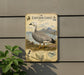 a metal plaque with an illustration of a white and black bird, likely an emu goose, standing on a grassy area with mountains in the background.