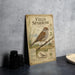 a vintage-style field sparrow illustration on a metal plate, placed on a table next to some black camera lenses.