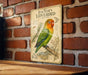 a vintage-style metal sign with a colorful illustration of a green and orange lovebird perched on a branch, set against a brick wall.