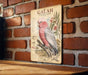 a vintage-style illustration of a pink cockatoo perched on a branch, with a brick wall in the background.