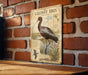 a vintage-style metal plaque with an illustration of a bird, likely a stork, standing on a brick wall.