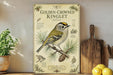 a vintage-style poster or sign with illustrations and text about the Golden-Crowned Kinglet, a small bird with a yellow head and green body, perched on a branch with pine needles. The poster is displayed on a shelf next to a wooden cutting board and a potted plant, creating a rustic and nature-inspired setting.