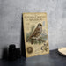 a vintage-style metal plaque with an illustration of a golden-crowned sparrow perched on a branch, surrounded by pine needles and pine cones. The plaque is placed on a gray countertop, with a stack of black camera lenses and a white plate visible in the background.