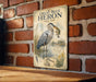 A vintage-style metal sign with an illustration of a great blue heron holding a fish, mounted on a brick wall.
