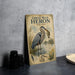 a framed illustration of a great blue heron with a fish in its beak, standing on a table next to a stack of black camera lenses.