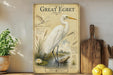 A vintage-style illustration of a great egret, a large wading bird, standing on a log in a pond with a lotus flower nearby. The illustration is framed by a decorative border and is displayed on a shelf alongside a wooden cutting board and a potted plant.